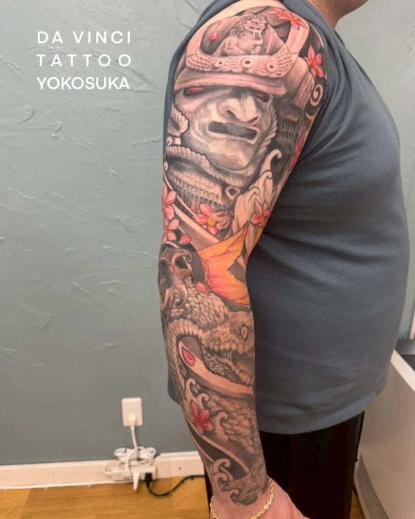 Person shown from the side wearing gray shirt. On his arm to shoulder large detailed traditional Japanese tattoo. Sleeve tattoo features samurai helmet, snake, scattered cherry blossoms, waves and red-orange koi fish.