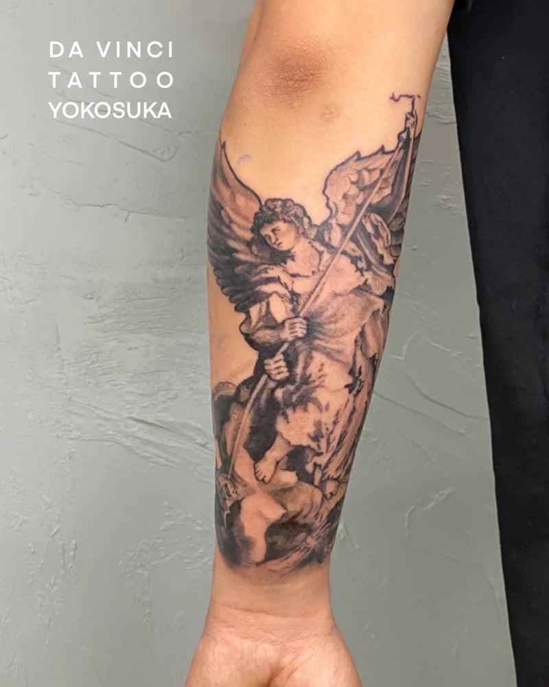 A close-up of a person's forearm featuring a large black and gray tattoo of an angel with wings and a spear pointed towards the fallen angel. against a light gray textured wall. White text in the upper left reads 'DA VINCI TATTOO YOKOSUKA'.