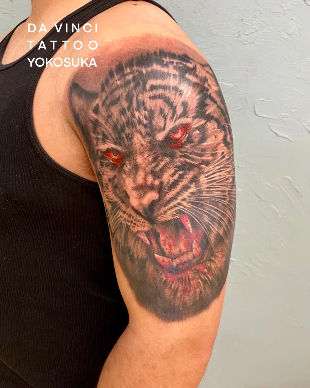 Da Vinci Tattoo Yokosuka black and gray realism. A realistic 3D-effect tattoo of a roaring tiger on a person's upper arm, with red eyes and an open mouth showing teeth, fine details, light gray wall background.