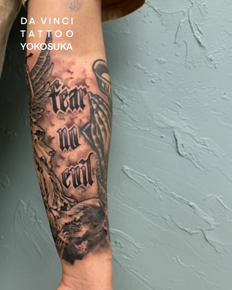 A close-up of a person's forearm featuring a detailed black and gray tattoo with the words 'Fear and evil' in gothic lettering, alongside winged and prayer hand motifs. The background is a textured light blue-gray wall, and white text in the upper left reads 'DA VINCI TATTOO YOKOSUKA'.