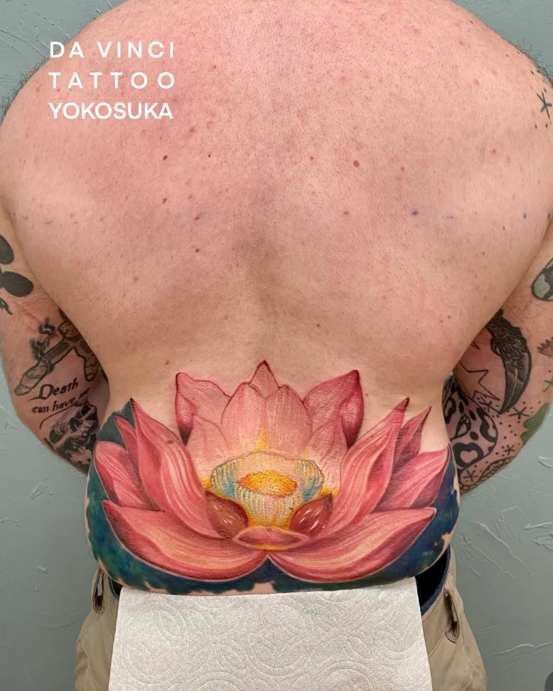 Da Vinci Tattoo Yokosuka. A close-up of a person's bare back showing a large, vibrant colorful lotus flower tattoo on the lower back. The tattoo features pink and red petals with a yellow stamen and dark blue-green background.