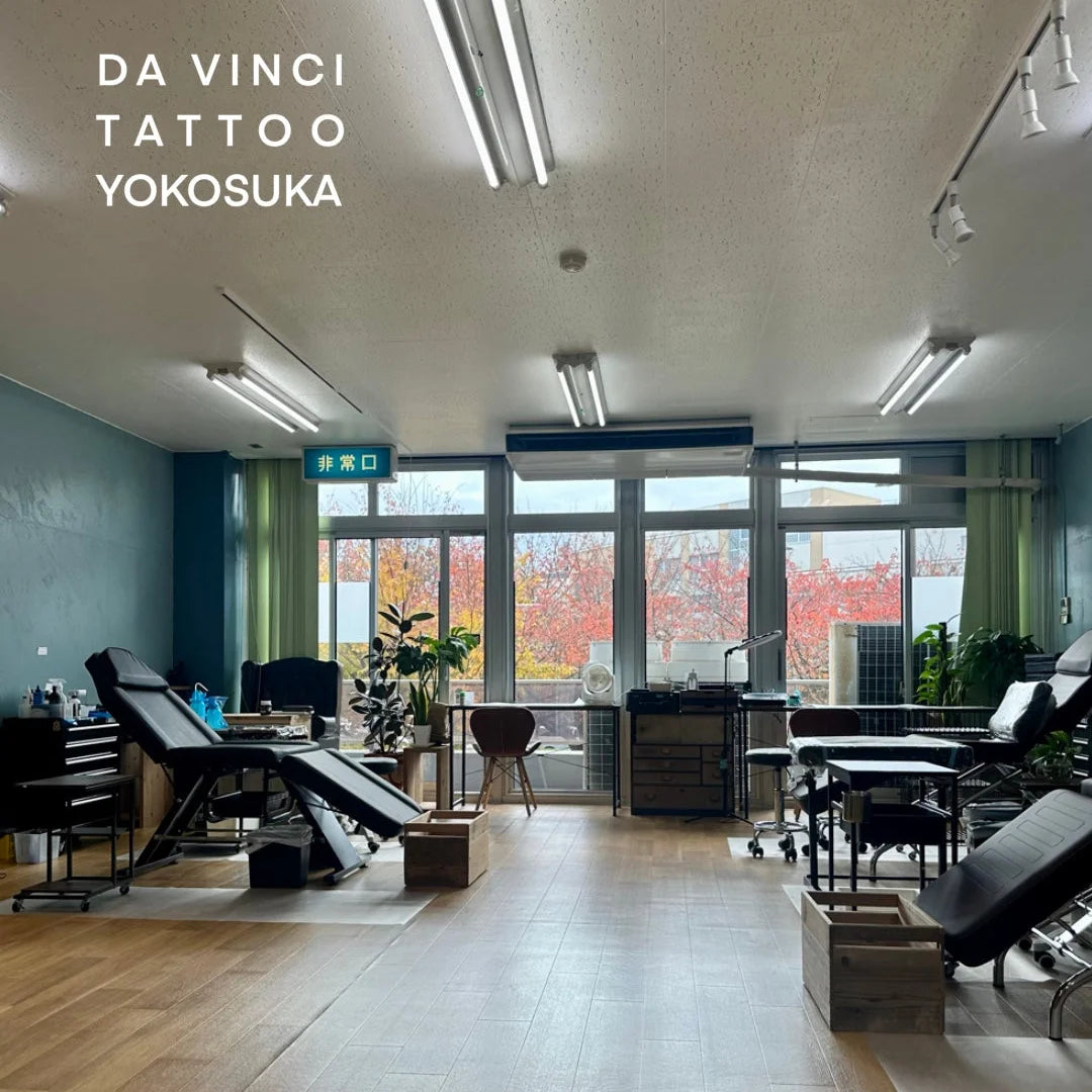 A spacious clean tattoo studio interior with professional black tattoo beds, work tables, and equipment arranged in a bright room with autumn window view and wood flooring. White text in the upper left reads 'DA VINCI TATTOO YOKOSUKA'.