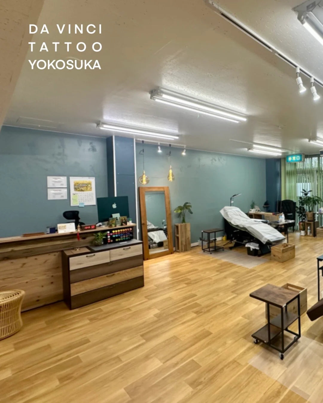 A tattoo studio interior with light wood flooring, teal walls, a reception desk, clean tattoo stations and bed, and the text 'DA VINCI TATTOO YOKOSUKA' in the upper left.