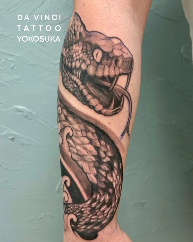 A realistic black and gray snake tattoo on a person's forearm, with the text 'DA VINCI TATTOO YOKOSUKA' in the upper left corner against a textured light green wall.