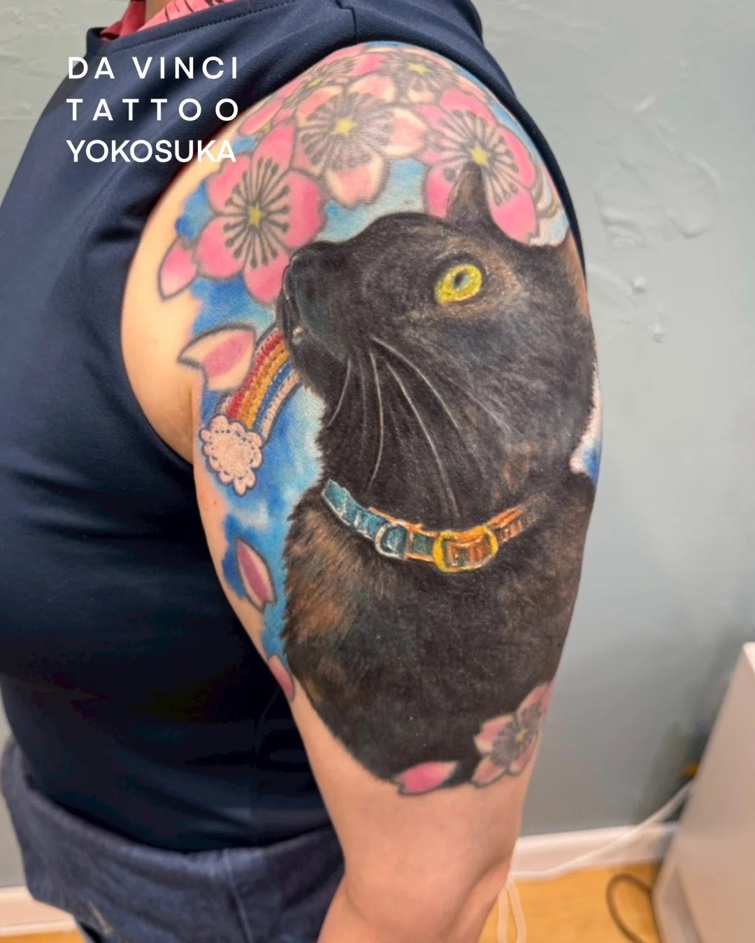 A large, colorful tattoo on a person's upper arm featuring a black cat with yellow eyes, pink flowers, and blue background details. The person is wearing a dark navy sleeveless top, and the text 'DA VINCI TATTOO YOKOSUKA' is visible in the top left corner.