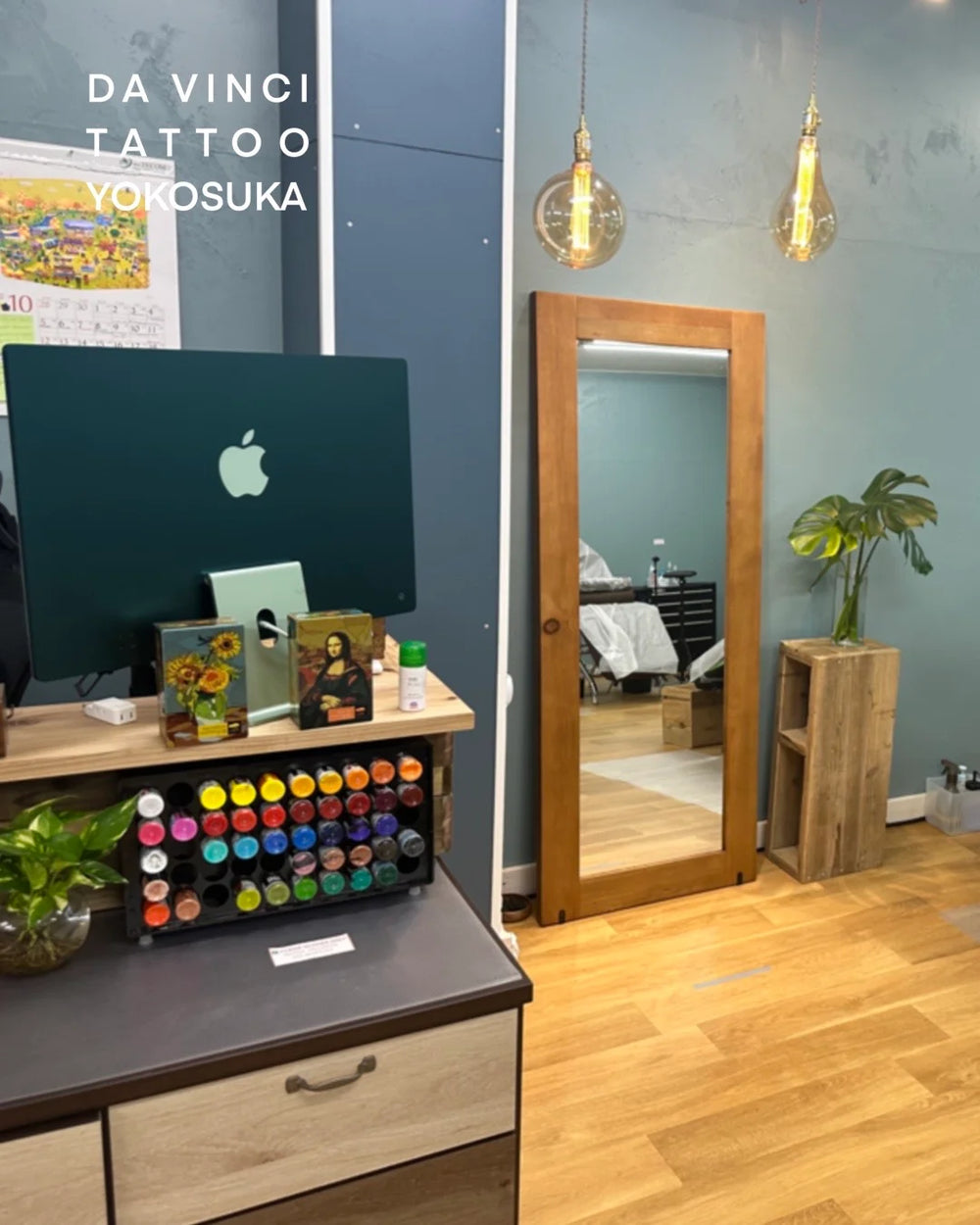 An interior view of 'Da Vinci Tattoo Yokosuka' clean studio featuring a wooden-framed full-length mirror, a desk with an Apple iMac, a colorful array of tattoo ink bottles, and warm hanging pendant lights against a blue-gray wall.
