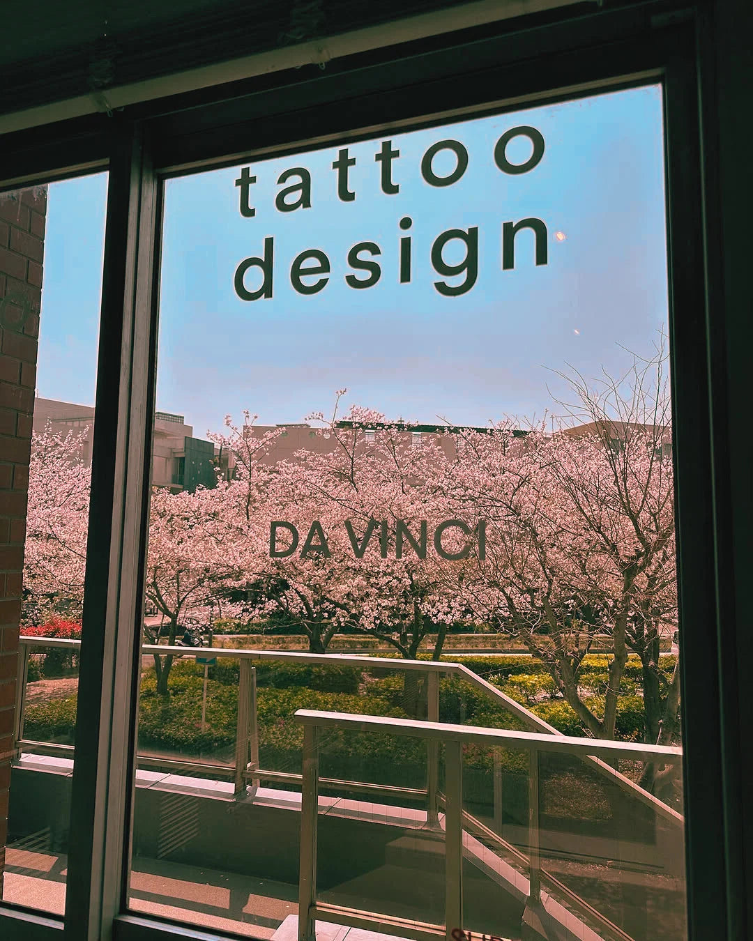 A view through a large window with the words 'tattoo design' and 'DA VINCI' printed on it, showing an outdoor scene with blooming pink trees and buildings under a blue sky.